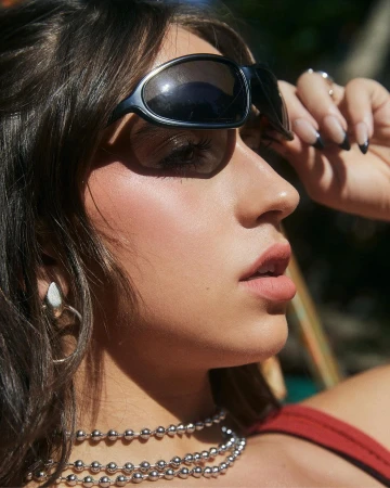 Close-up portrait of a woman wearing dark wraparound sunglasses and layered silver necklaces, captured by Sami Drasin.