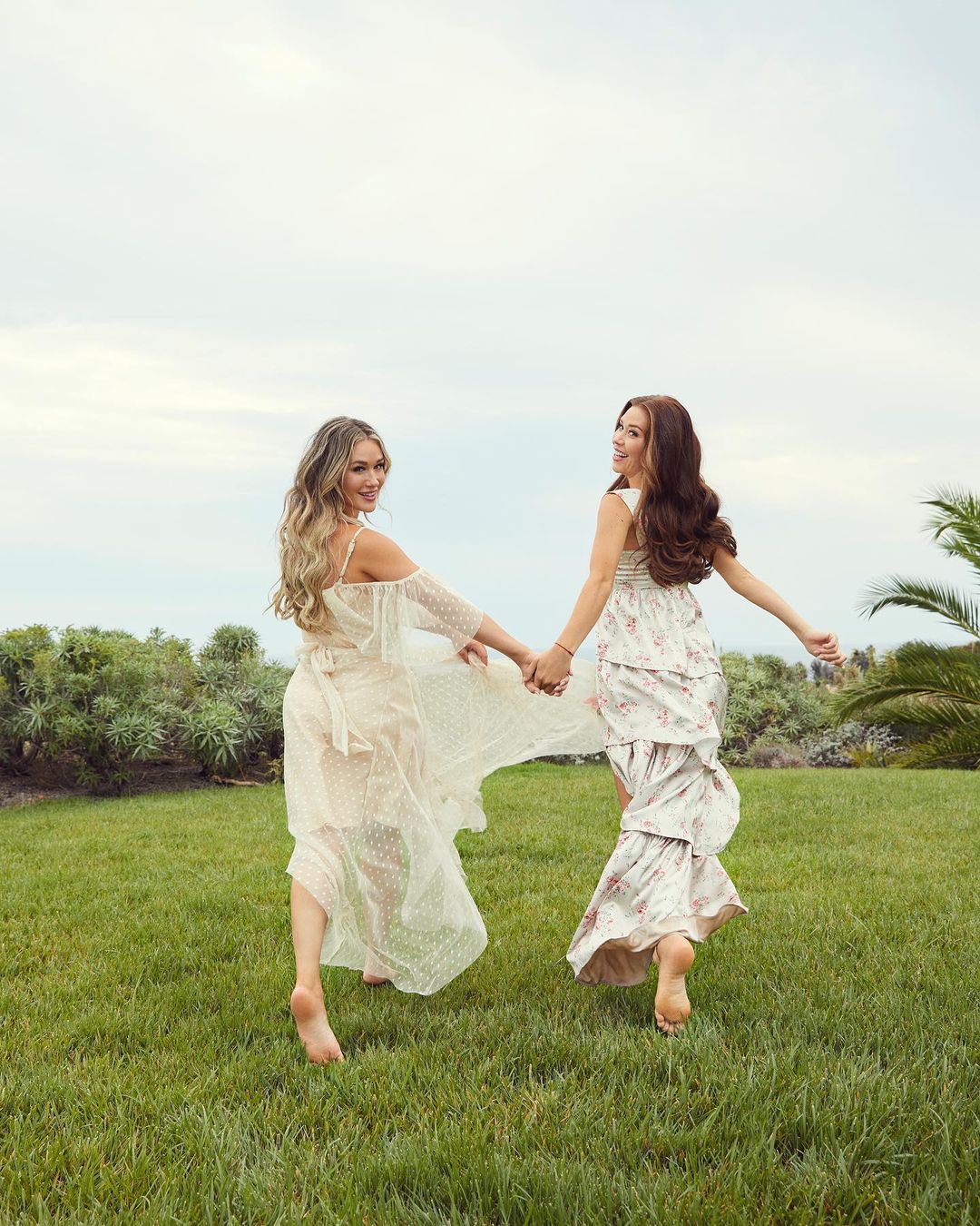 Two barefoot women in long floral dresses hold hands, strolling through green grass with trees and cloudy sky in the background.