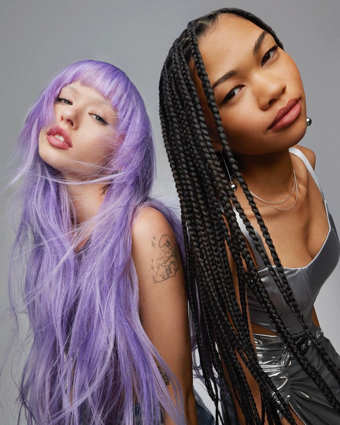 Two women pose closely together in a studio shot. One has long, braided black hair, and the other has long, wavy lavender hair.