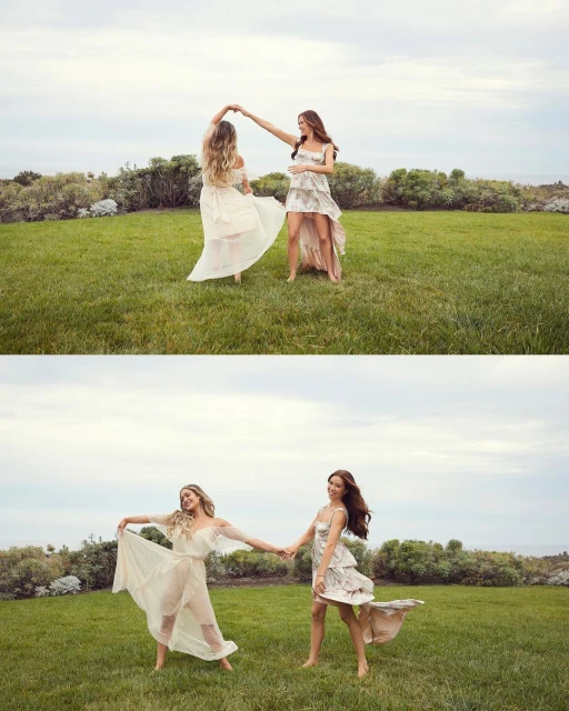 Two women in dresses twirl on a green lawn with coastal vegetation and sky in the background, captured in an eye-level medium shot.