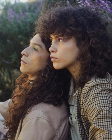 Two people with long curly brown hair and patterned jackets posing closely together outdoors near purple flowers.