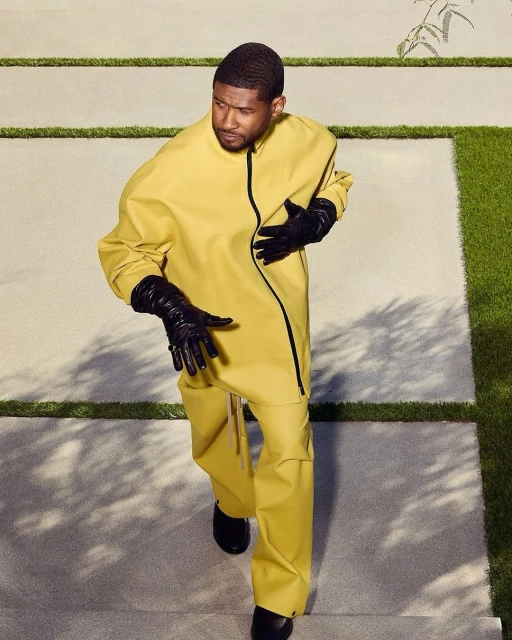 Usher poses on concrete steps in yellow outfit and black gloves in an eye-level medium shot with natural lighting and green foliage.
