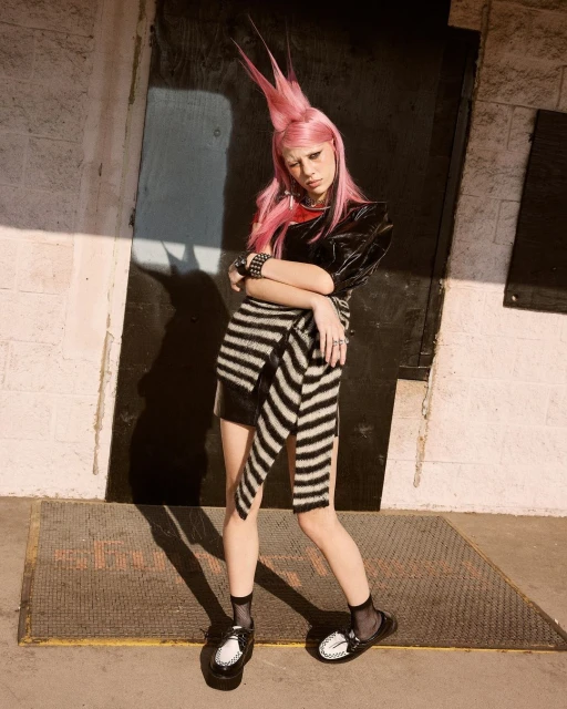 A slender person with pink spiky hair poses outdoors in a dark top, striped wrap skirt, and shiny loafers in front of a dark doorway.