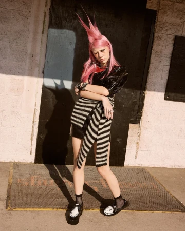 A slender person with pink spiky hair poses outdoors in a dark top, striped wrap skirt, and shiny loafers in front of a dark doorway.