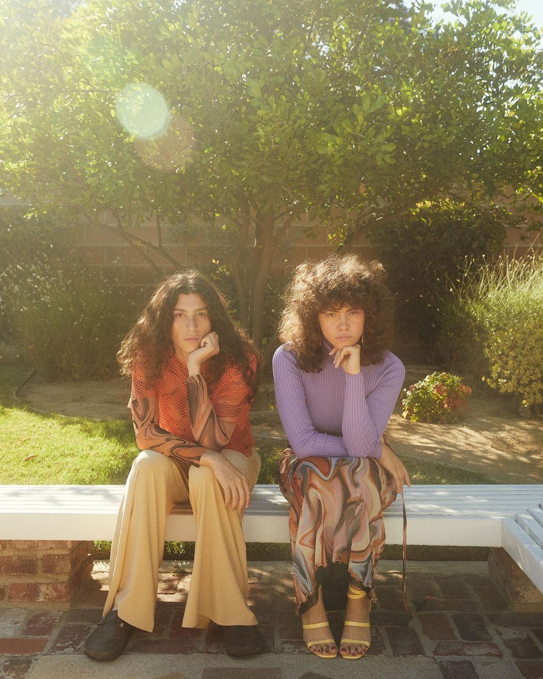 Two women sit side-by-side outdoors on a white bench surrounded by lush green trees and wearing retro-style clothing in a sunlit setting.