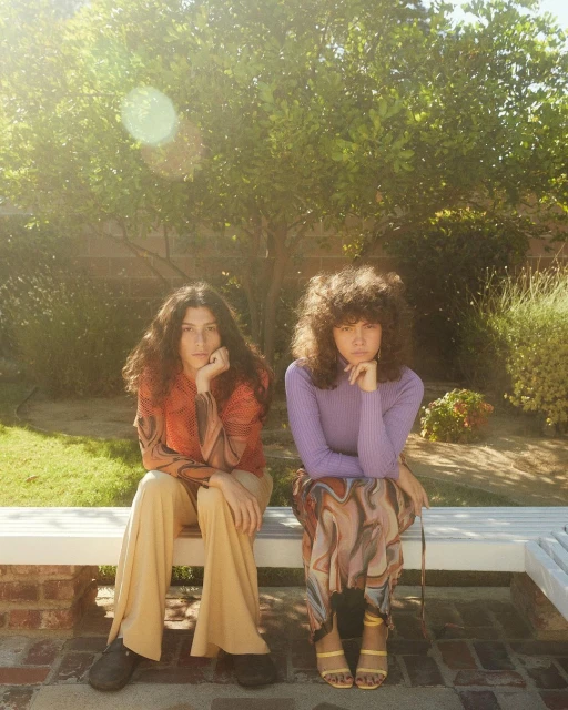 Two women sit side-by-side outdoors on a white bench surrounded by lush green trees and wearing retro-style clothing in a sunlit setting.