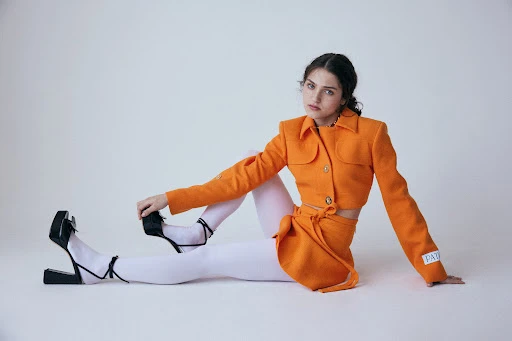 Brunette woman in orange blazer and skirt, sitting, and black heels, against white backdrop. Fashion editorial style, medium shot.