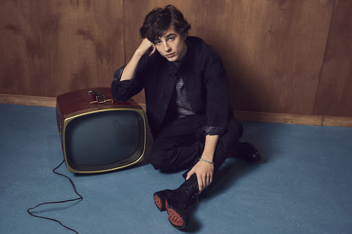 A young adult male, seated next to a vintage television, rests his head on his hand while looking toward the camera indoors.