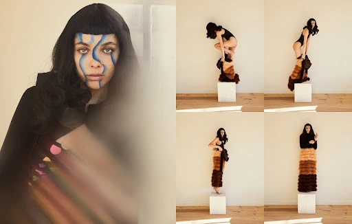 Surreal fashion portrait: A woman with face paint stands on a white block, wearing a brown fringed garment against a white wall backdrop.