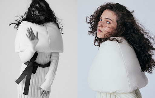 Woman with long curly dark hair wearing a white puffed sleeveless top over a ribbed dress cinched with a black belt