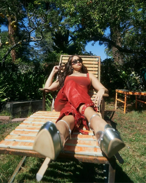 Woman lounges in an outdoor chair, wearing sunglasses, red outfit, and silver platform shoes, amidst lush greenery and dappled sunlight.