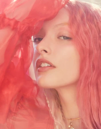 Close-up portrait of a woman with pink hair lifting a sheer red sleeve near her face, photographed by Sami Drasin.