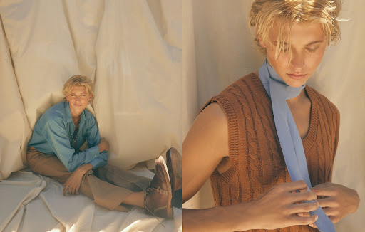 Young adult with blond hair poses in front of draped white fabric, wearing a blue shirt, brown vest, tan trousers and brown shoes.