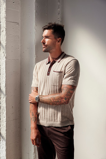 Man with tattoos wearing a ribbed knit polo shirt and dark trousers standing indoors by a wall