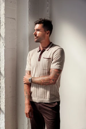 Man with tattoos wearing a ribbed knit polo shirt and dark trousers standing indoors by a wall