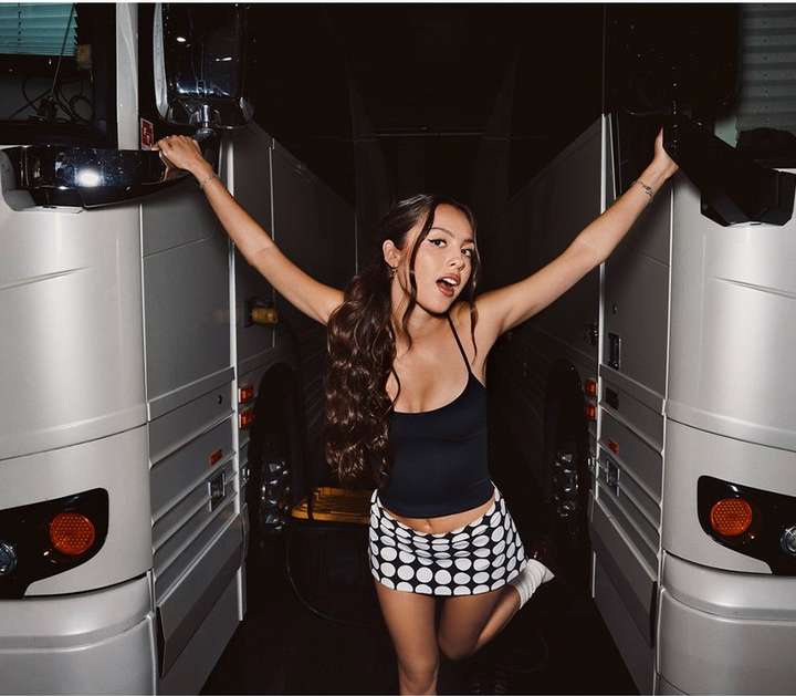 A young adult female with long brown hair in a black top and polka dot skirt poses in front of two tour buses, smiling with arms raised.