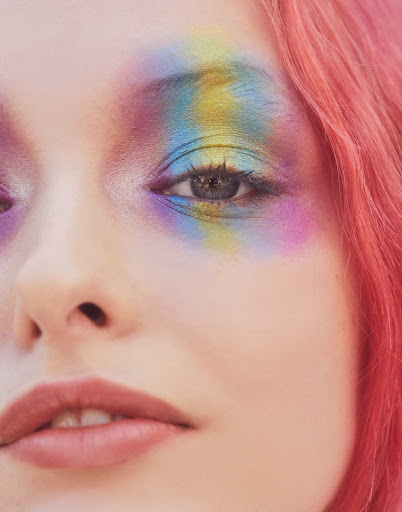 Close-up of a person's face with pink hair and rainbow eye makeup, capturing a striking and colourful beauty portrait.