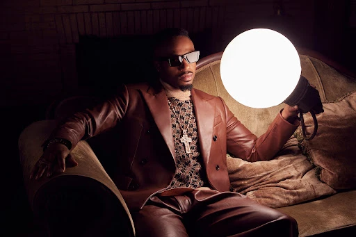 Portrait of an adult male in a brown leather suit sitting on an ornate couch holding a large glowing orb in a dimly lit indoor setting.