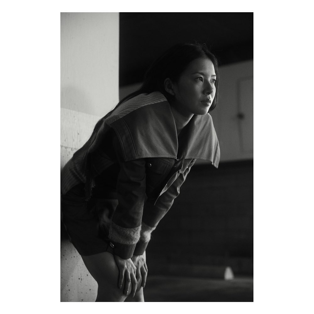 Portrait of an Asian woman with hands resting on her knees, looking away from the camera, wearing a casual jacket in monochrome lighting.