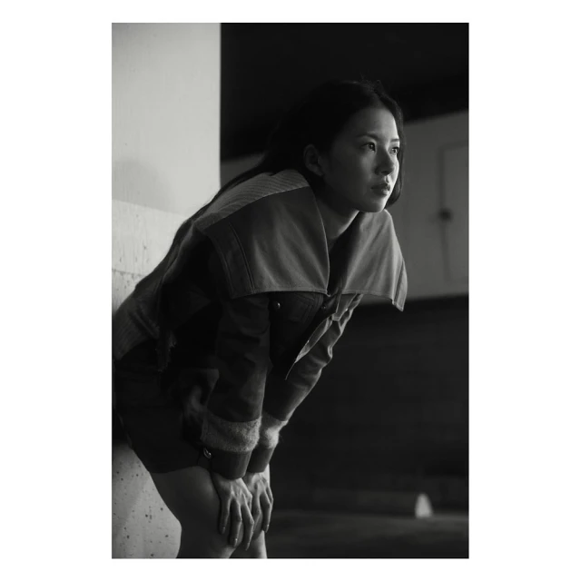 Portrait of an Asian woman with hands resting on her knees, looking away from the camera, wearing a casual jacket in monochrome lighting.