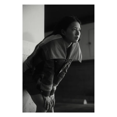 Portrait of an Asian woman with hands resting on her knees, looking away from the camera, wearing a casual jacket in monochrome lighting.