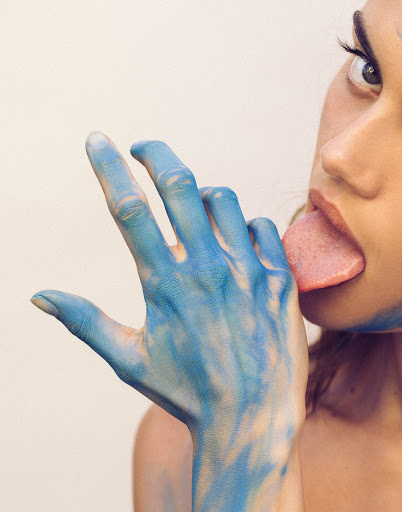 Close-up portrait of a woman with blue paint on her hand and face, sticking out her tongue against a plain light background