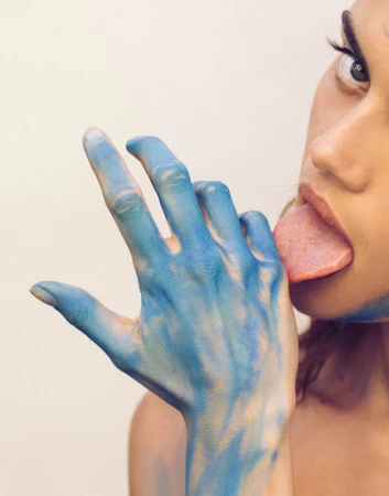 Close-up portrait of a woman with blue paint on her hand and face, sticking out her tongue against a plain light background