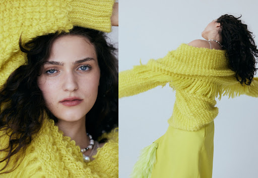 Brunette woman poses in a fuzzy yellow sweater and skirt against a pale backdrop, gazing upwards with arms raised in a fashion editorial.