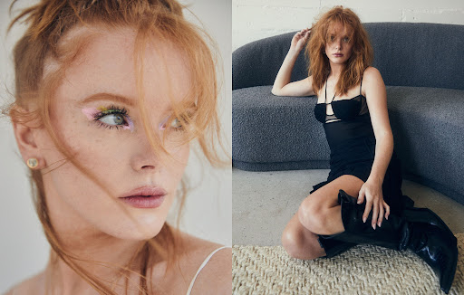 Portrait of a red-haired woman in a black dress and boots, posing on a beige carpet in front of a gray couch in an indoor setting.