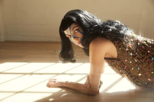Woman with dark hair, light skin, and face paint in brown patterned dress lying on light wood floor with window light reflecting on it.