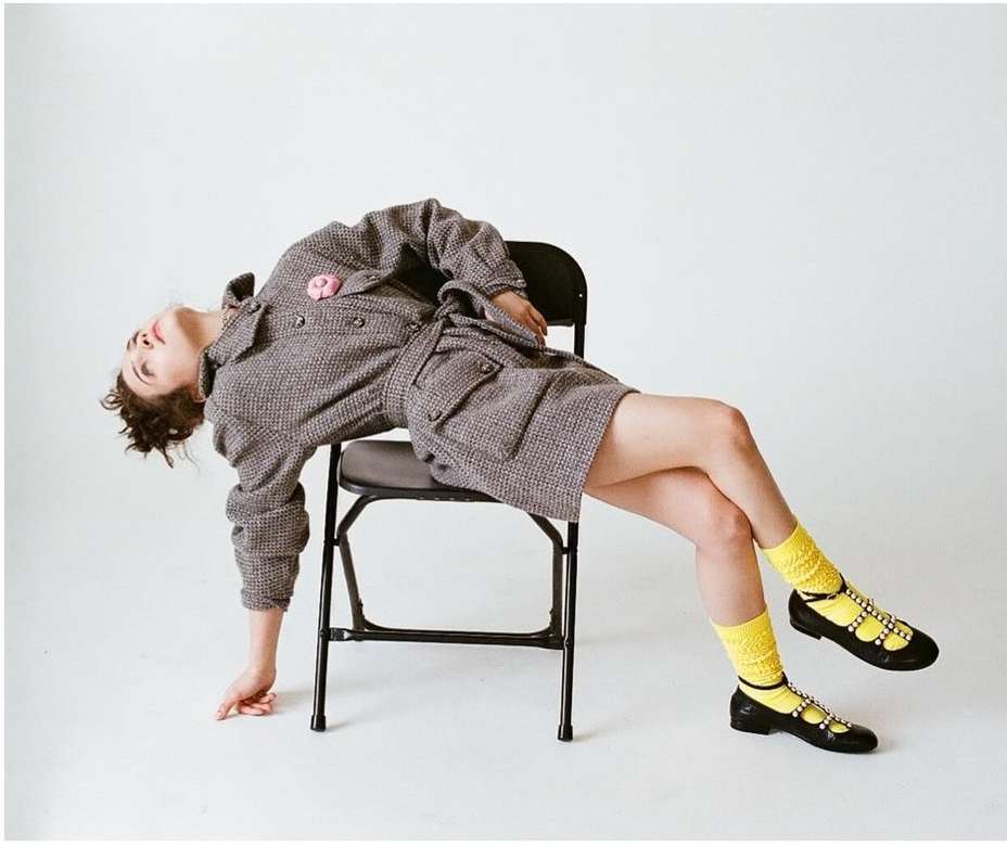 Woman in tweed dress and yellow socks posing on black folding chair in white studio. Fashion, portrait, and studio photography.