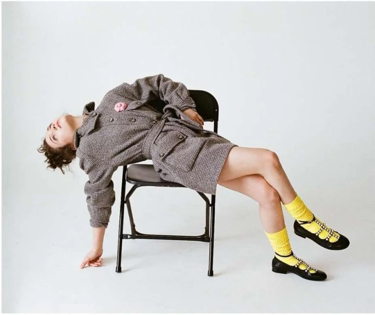 Woman in tweed dress and yellow socks posing on black folding chair in white studio. Fashion, portrait, and studio photography.