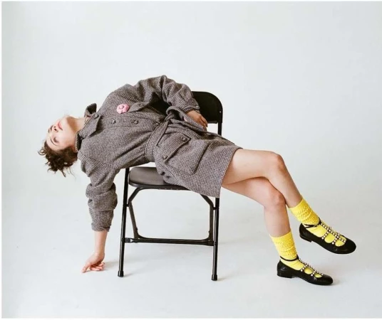 Woman in tweed dress and yellow socks posing on black folding chair in white studio. Fashion, portrait, and studio photography.