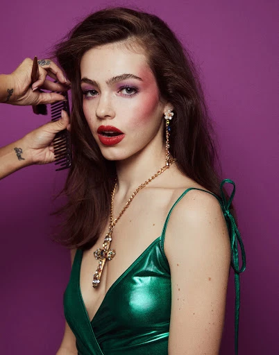 A woman with red lipstick poses with a green dress while a stylist adjusts her hair in a studio with a violet background.