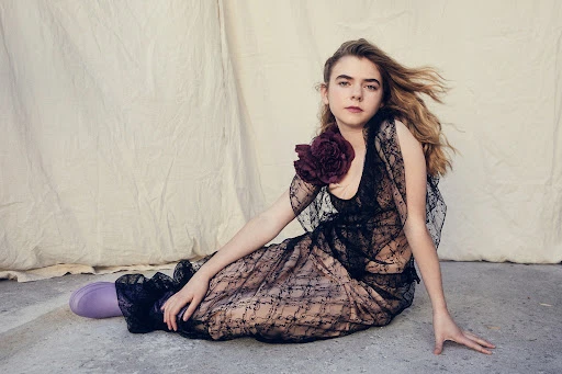 Young woman in a black lace dress with a purple flower brooch sitting on a concrete floor before a draped backdrop.