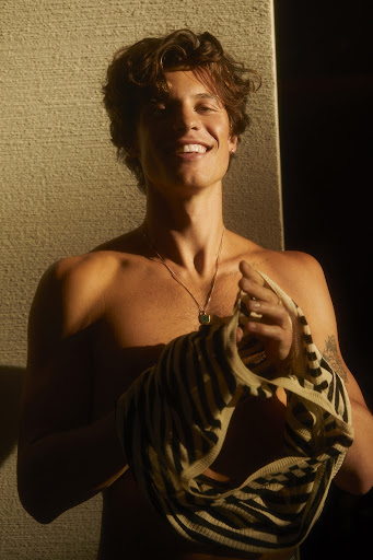 Smiling shirtless man with curly brown hair holding a striped top in a studio with light and shadow contrasts and a neutral color palette.