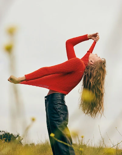 Woman in red sweater and black leather pants poses in a field of yellow wildflowers against an overcast sky, hair flowing.