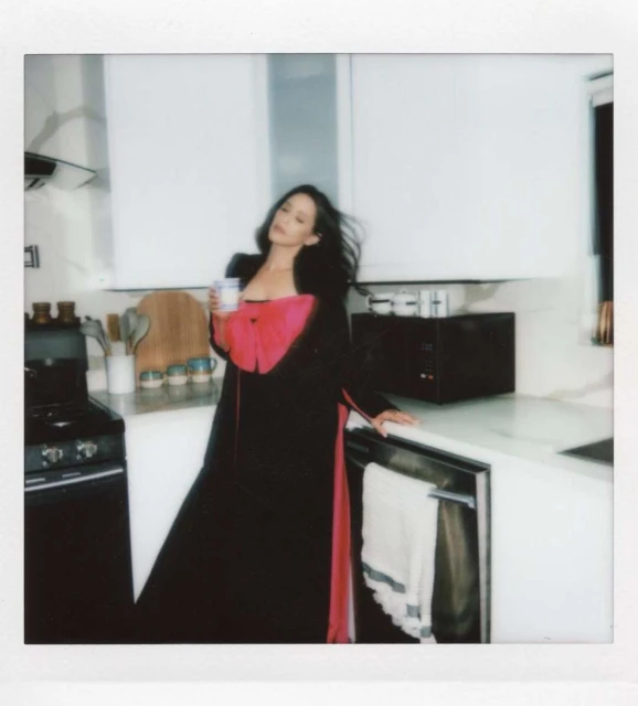 A woman in a black and red robe poses in a modern kitchen with white cabinets, holding a mug in a lifestyle shot. 