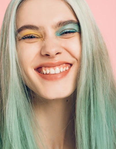 Close-up studio shot of a smiling young woman with ombre mint-green hair, colorful makeup, and pink lips against a pink background.