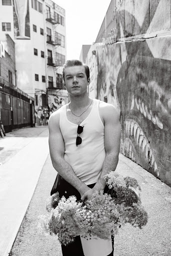 Black and white portrait of a young man in a tank top holding flowers in an alleyway with graffiti walls and a building in the background.