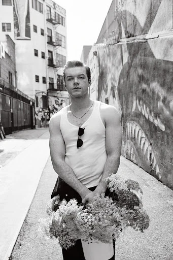 Black and white portrait of a young man in a tank top holding flowers in an alleyway with graffiti walls and a building in the background.