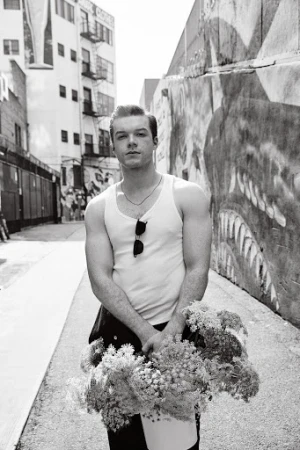 Black and white portrait of a young man in a tank top holding flowers in an alleyway with graffiti walls and a building in the background.