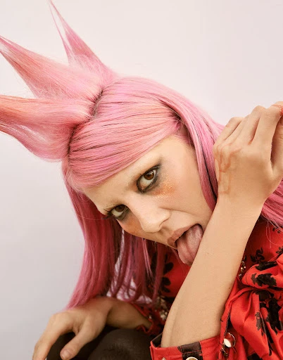 Unique portrait of a young woman with pink spiky hair and heavy makeup, sticking her tongue out, wearing a patterned top against white.