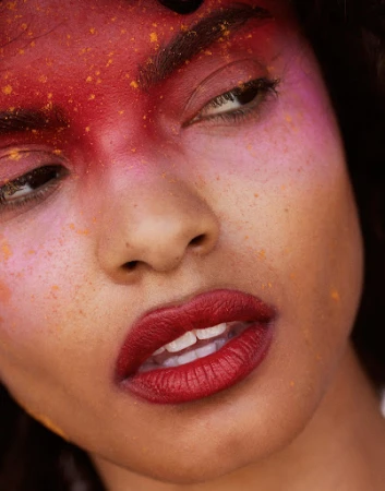 Close-up portrait of a woman with red lipstick and face paint in hues of red, pink, and orange, and a dark hair color.