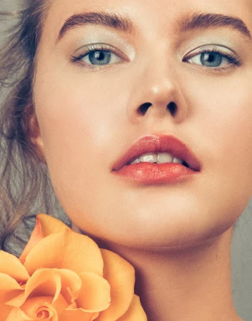 Close-up portrait of a fair-skinned woman with blue eyes and pink lips, alongside a yellow rose, showcasing beauty and skincare.