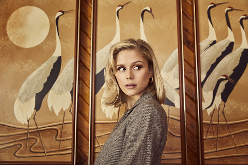 Portrait of a blonde woman in a grey blazer posing in front of three framed paintings of cranes in an indoor environment with soft lighting.