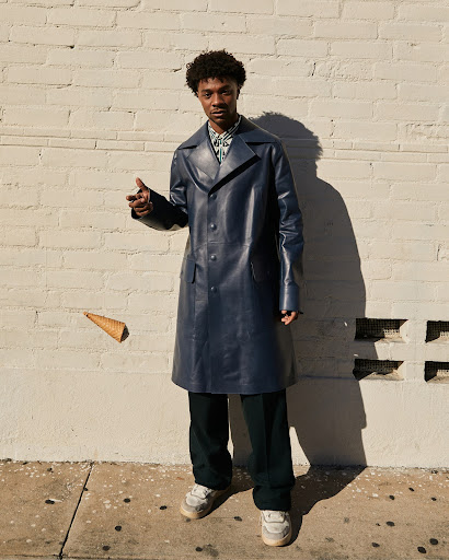 Stylish man in a blue trench coat poses outdoors next to a brick wall with a floating ice cream cone, creating a fashionable and playful image.