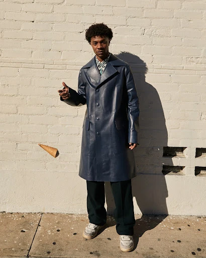 Stylish man in a blue trench coat poses outdoors next to a brick wall with a floating ice cream cone, creating a fashionable and playful image.
