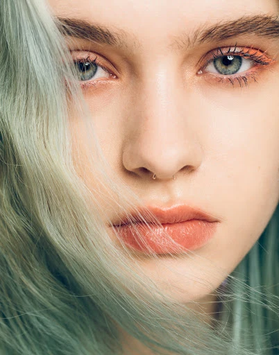 Close-up portrait of a fair-skinned young woman with green eyes, a nose ring, and light turquoise wavy hair framing her face.