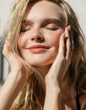 Close up portrait of a blonde woman with eyes closed holding her face with her hands in warm sunlight